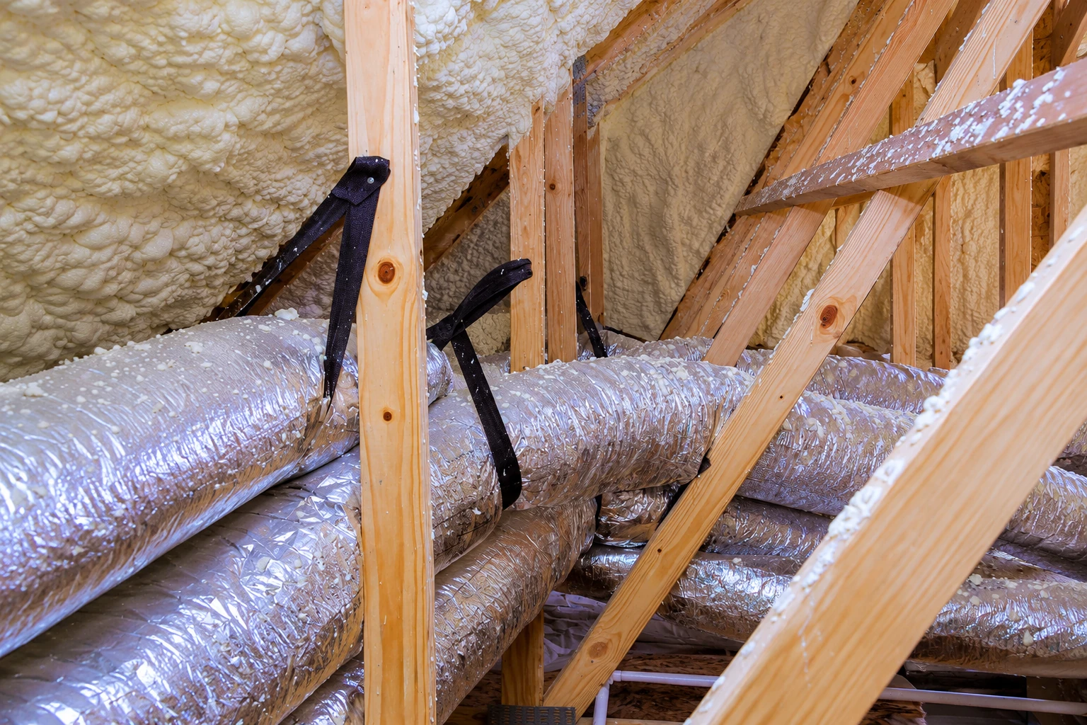 Foam insulation covers the roof framing above several silver flexible ducts in a wooden attic.