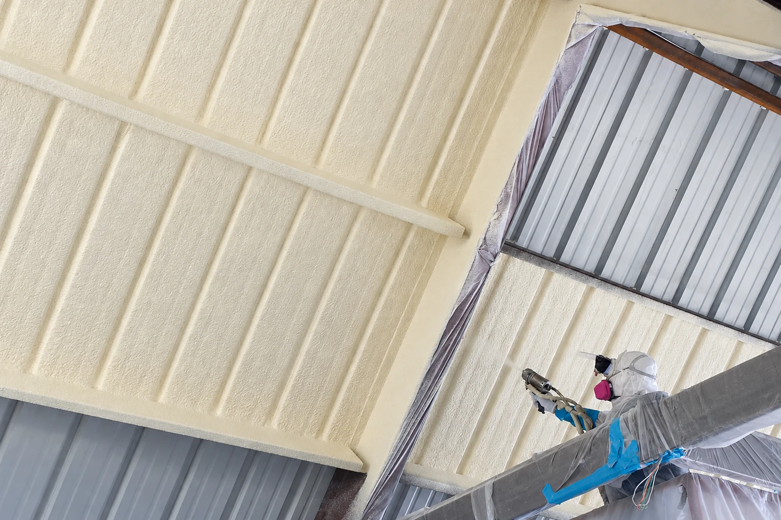 A worker in protective gear sprays pale foam insulation onto the corrugated metal roof of a commercial building.