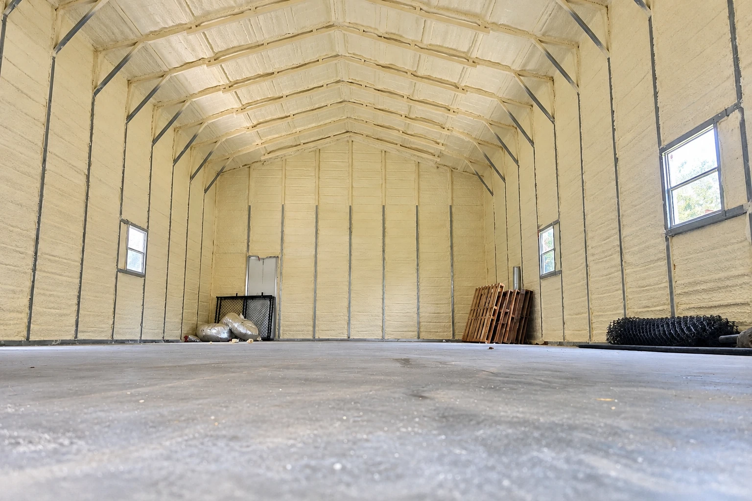 A large commercial building interior has pale spray foam coating its arched ceiling and walls, with a concrete floor and a few stored materials.