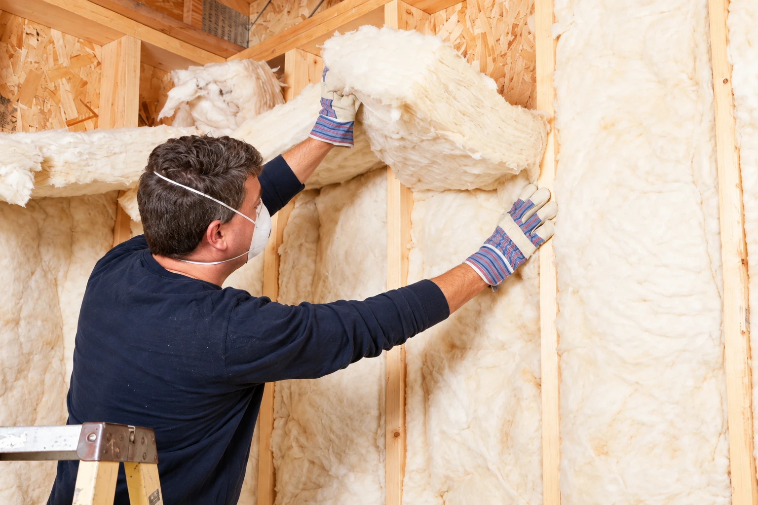 A masked worker in gloves fits fluffy fiberglass insulation between the studs of a wooden wall.
