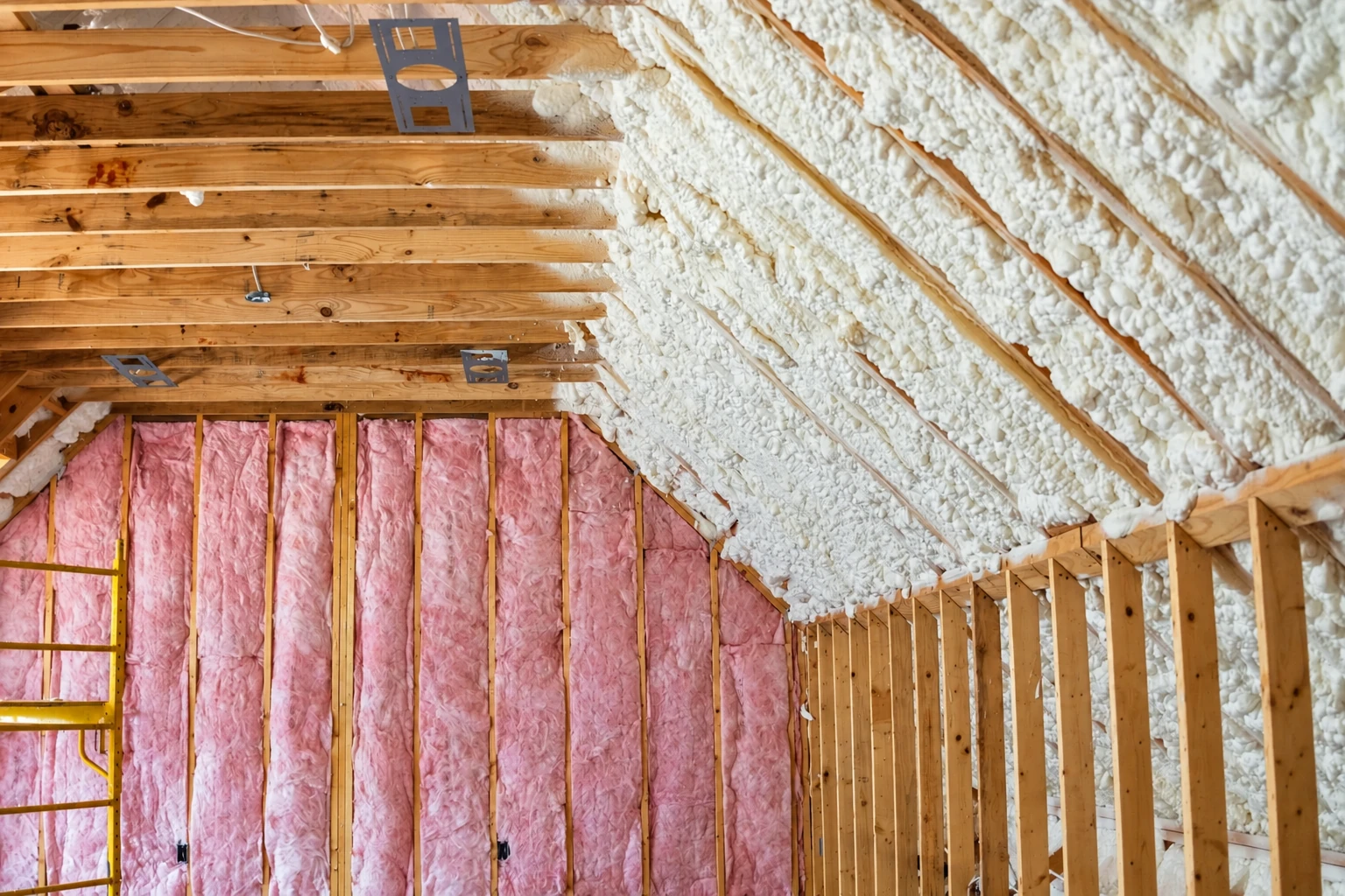 Attic rafters and sloped roof covered in white spray foam, above wall cavities filled with pink fiberglass batts.