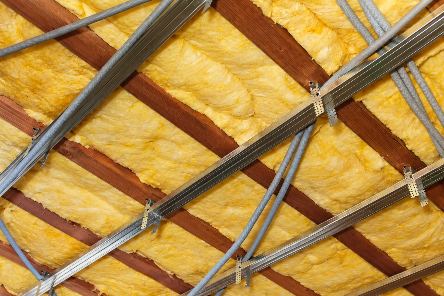 Yellow fiberglass insulation fills wooden ceiling joists crossed by metal framing and gray flexible conduits.