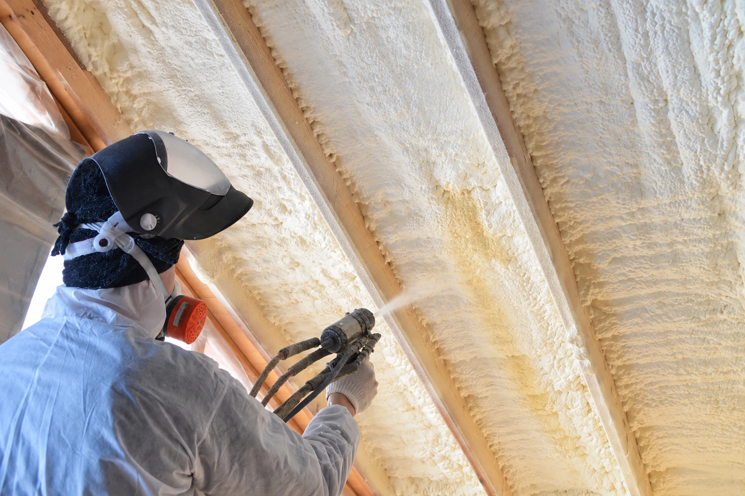 A worker in protective gear sprays foam insulation into exposed roof framing.