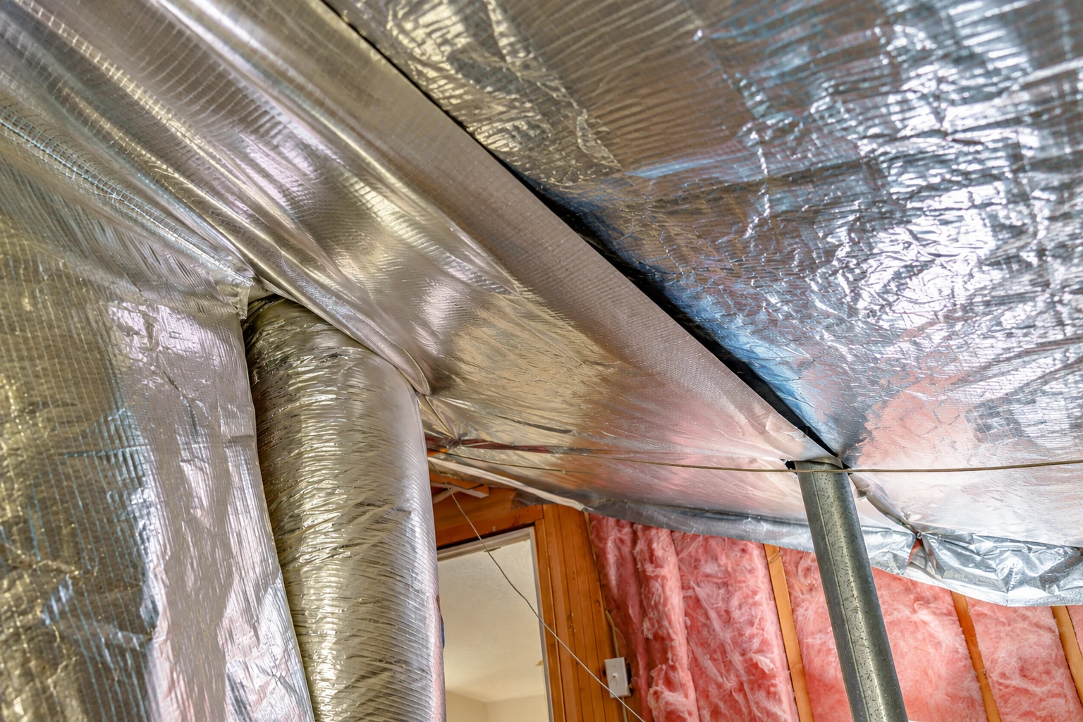 Reflective foil vapor barrier covers a ceiling around ductwork, with exposed pink insulation visible between wall studs.