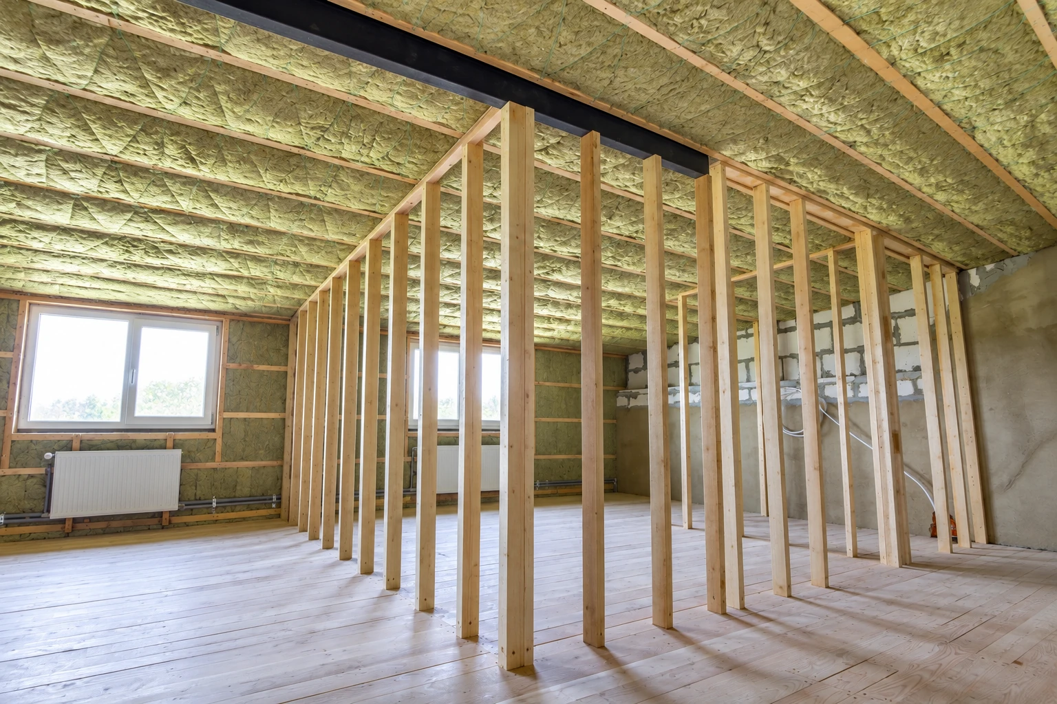 Unfinished room with green insulation between ceiling joists and wall studs, with exposed framing and a white-framed window.